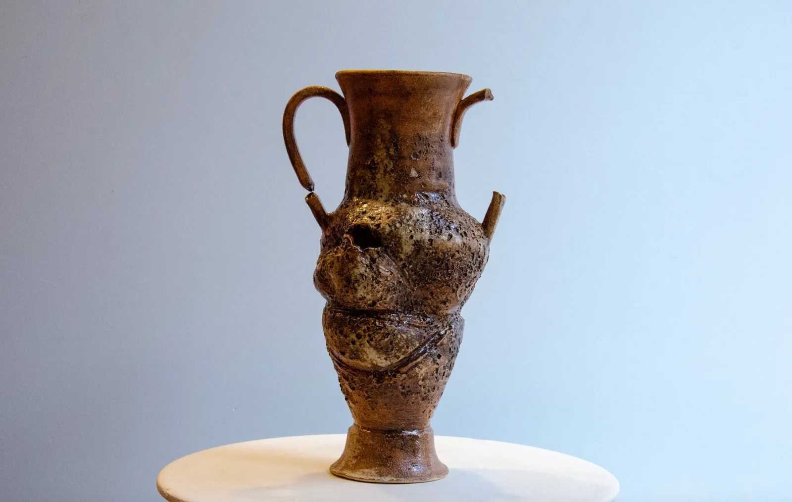 Photo of a finished amphora on a small, round surface. The handles are broken and missing in some places, and the body of the amphora is distorted with holes and texture.