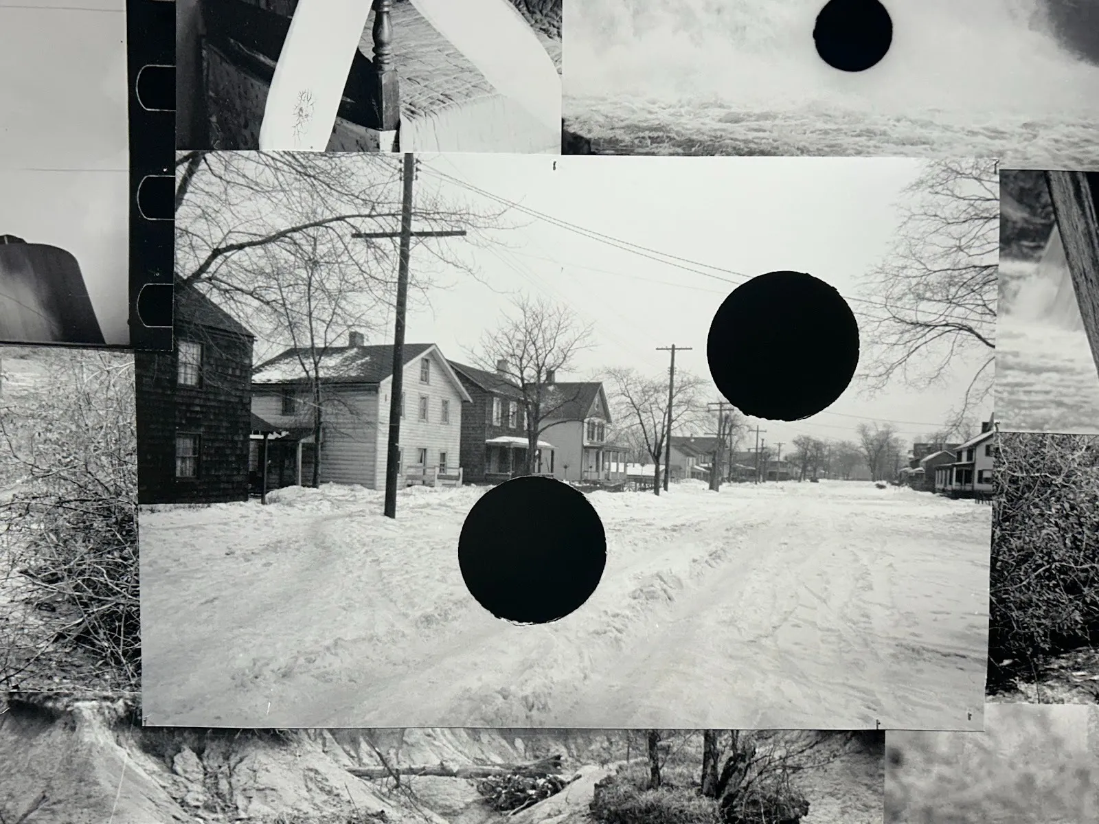 Detail shot of a central photograph laid on top of other black-and-white photos. The central photo is black and white, featuring a snowy residential street, with two black holes cut out.