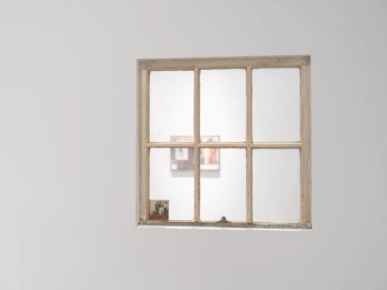Close-up shot of a white wall with a six-pane window opening.  The painted pane has a small photograph tucked into the bottom. Through the window, another room featuring a collage work can be seen.