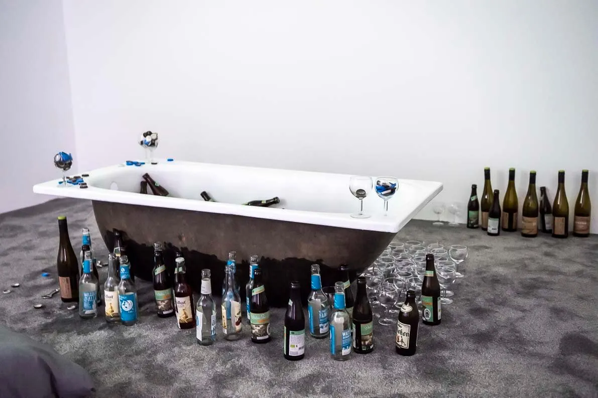 Still life, bathtub with beer bottles, wine bottles, glasses, and sparkling water bottles around it on grey carpet, Nikima Jagudajev, basically at westfallischer kunstverein.