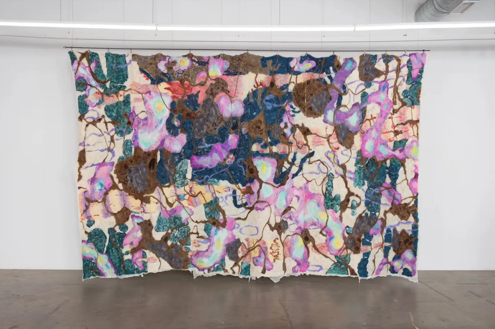 A large, colorful textile work hung from the ceiling features dark and light shades of purples, browns, and blues. The weaving forms take organic, nonlinear shapes.