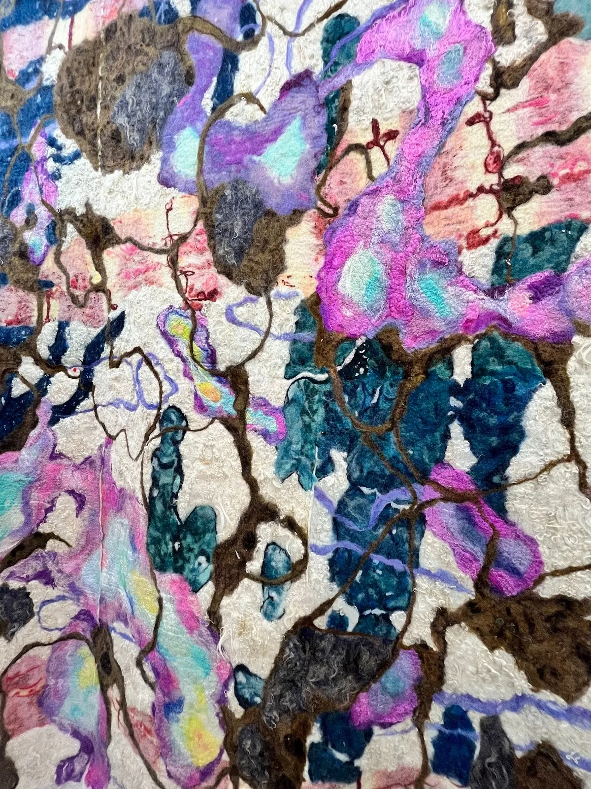 A detail shot of a flat textile work. Small fibers stick out slightly and have a matted, felted texture. Brown and iridescent-like purples and blues cross and weave over an off-white background.