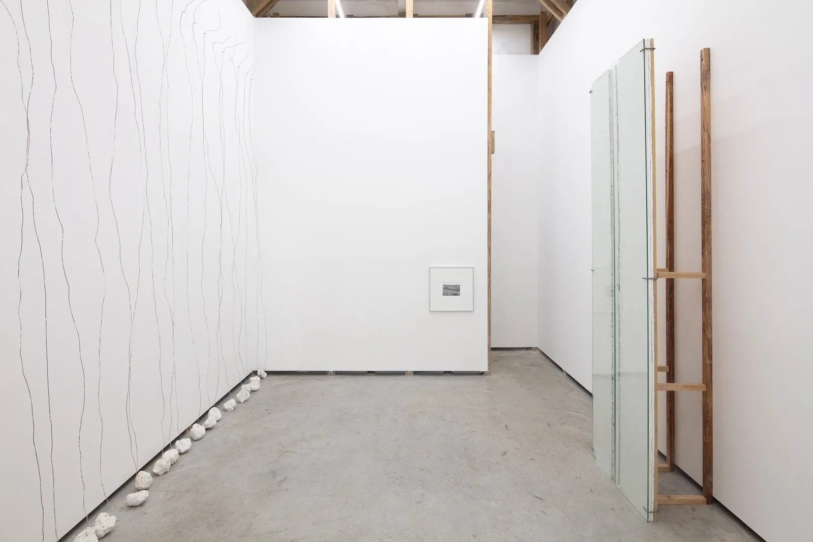 Constructed temporary walls stand in front of white gallery walls. The right wall has wires hanging down and weighed by rock-like shapes. The back wall has a low, small square B&W photo. The right wall has a pane of glass flush to its surface.