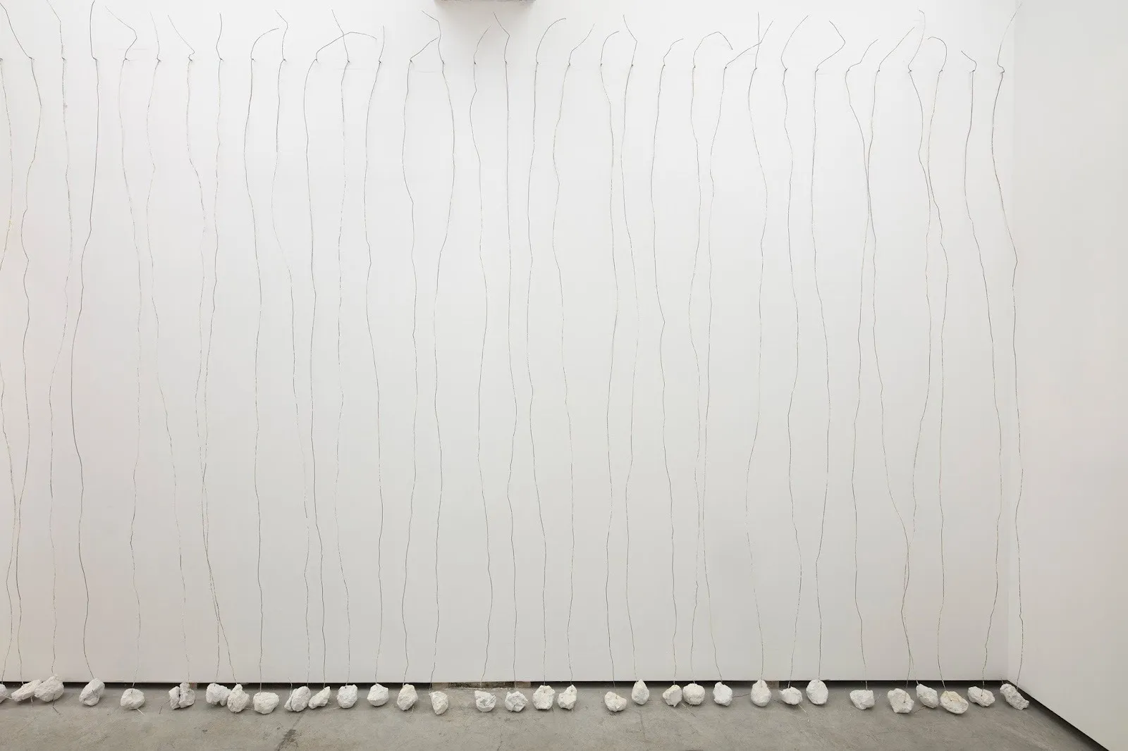 Shot of gallery with concrete floor. Two temporary white walls meet. Wires nailed to the upper part of the wall hang down and are weighed down by white rock-like objects.