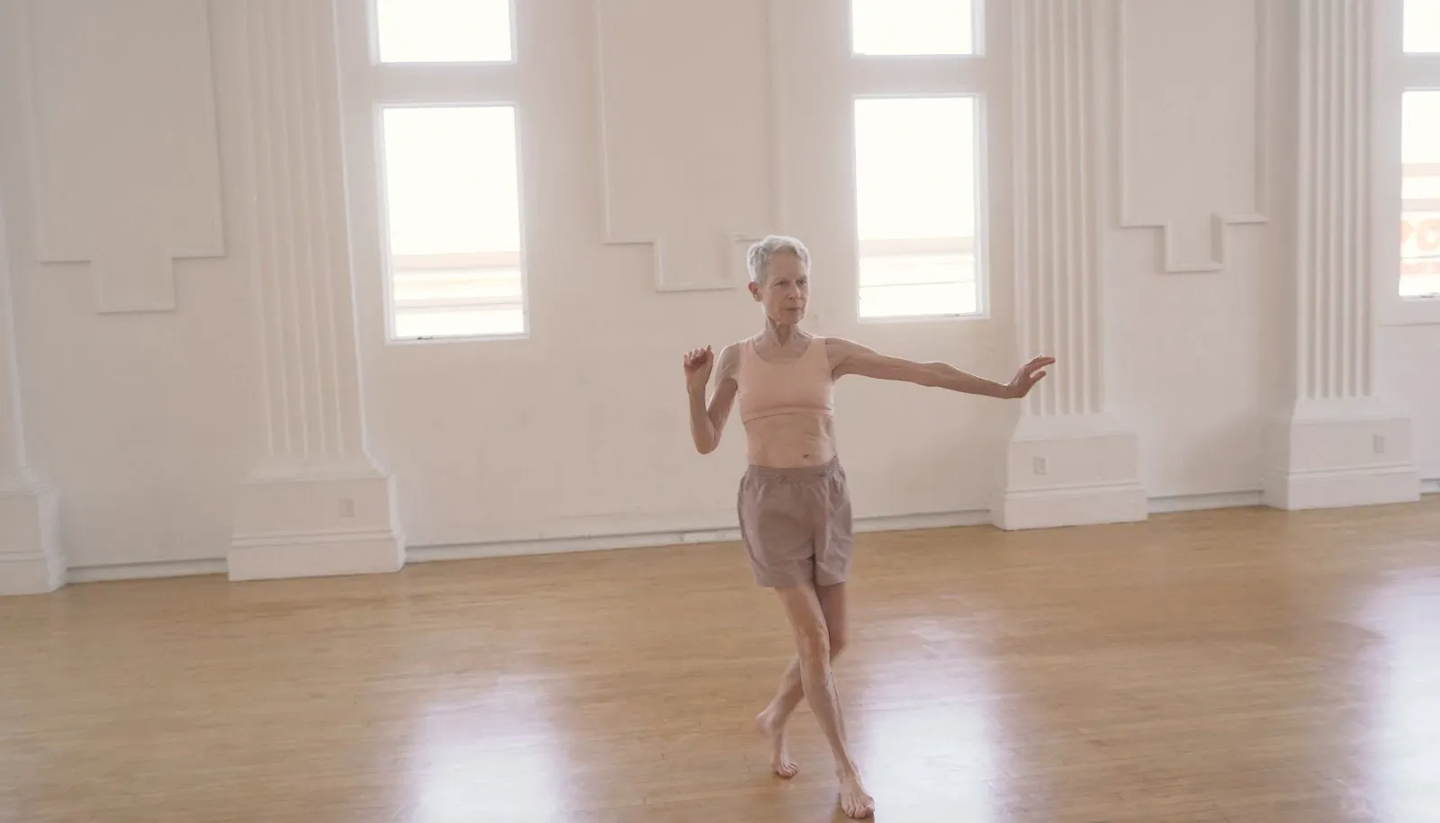A figure clad in neutral colors seems to dance, caught in mid-movement with one arm bent upwards and the other extended to her side. The figure dances in a white room with windows and a light hardwood floor.