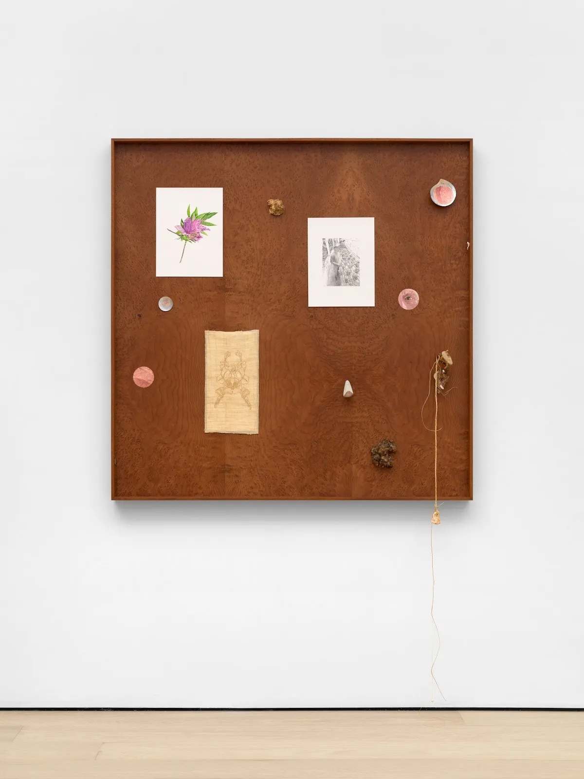 Frontal image of a darker, square wooden panel mounted to a white wall. Various media and images are placed on the panel, including a painting of a flower, embroidery work, and a b&w drawing. A piece of string nailed on the panel brushes the floor.