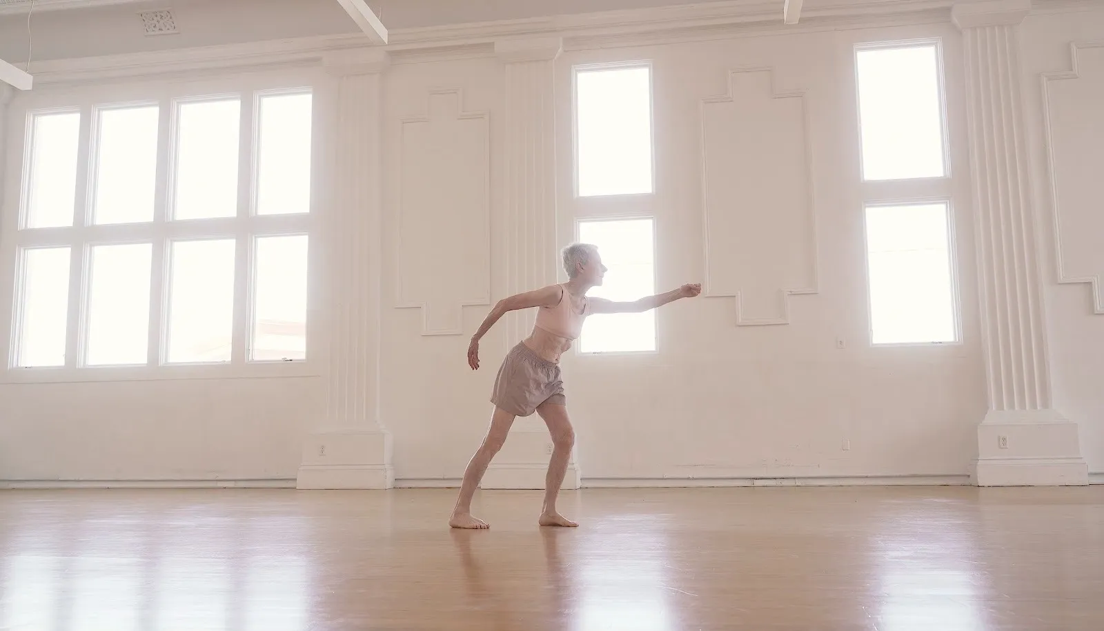 A figure clad in neutral colors seems to dance, caught in mid-movement. She seems to move to the right, leaning, one arm outstretched in front of her and another behind. The figure dances in a white room with windows and a light hardwood floor.