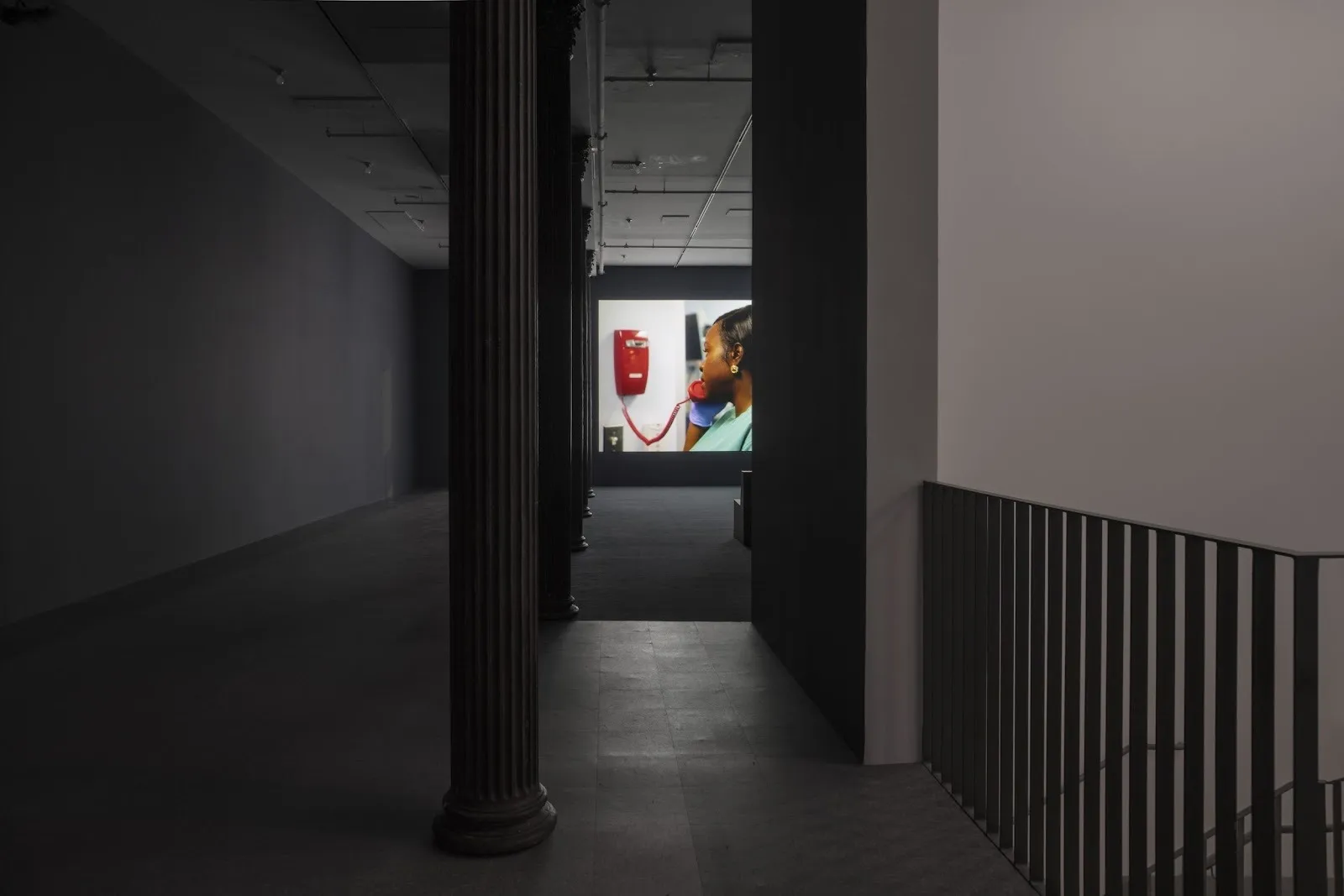Shot of dimly-lit gallery room with fluted colonnade. In the back of the shot, a wall projection features a woman mid-call on a red corded phone.