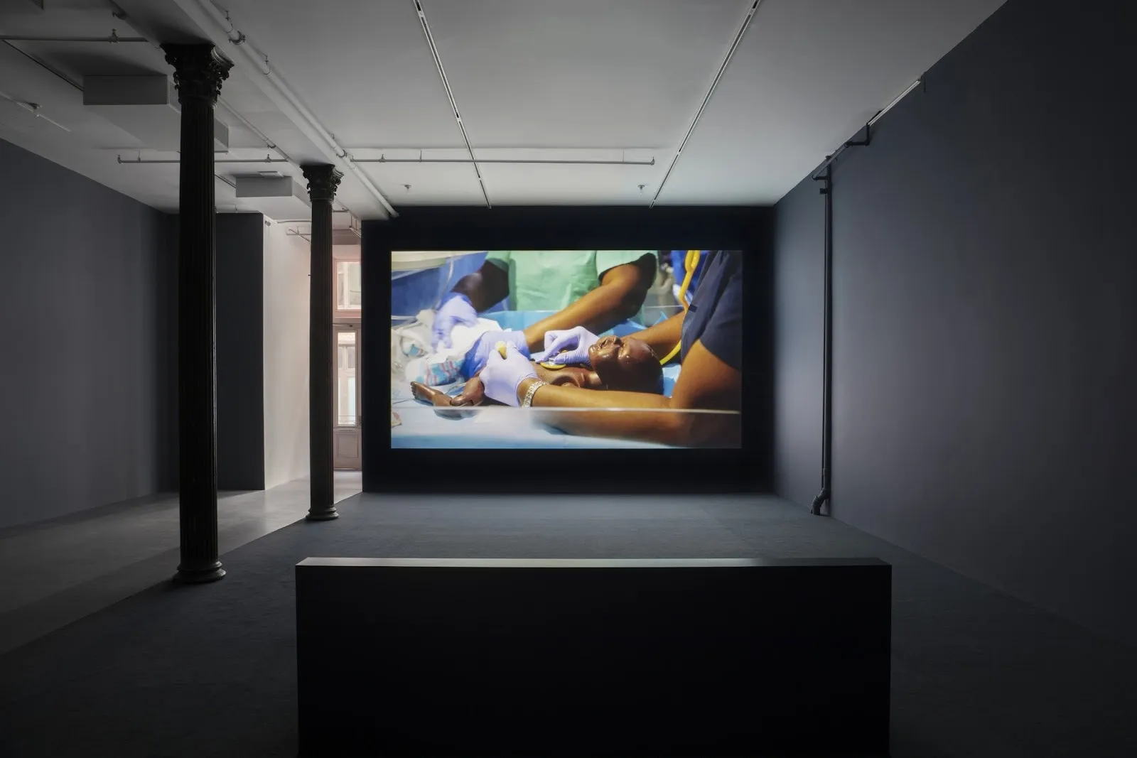 Dimly-lit gallery room with grey floors and fluted columns. Doorway in the far back. The wall facing the camera features a large projected video. Video depicts people in scrubs tending to a mannequin baby.