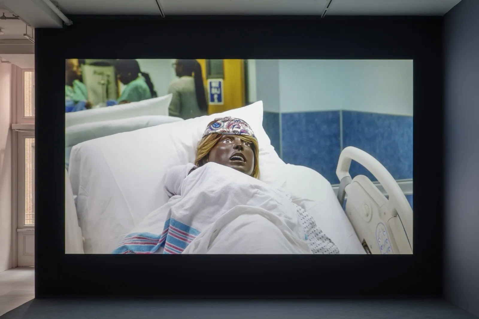 Video projection featuring mannequin nestled in blankets in a hospital bed. In the background, there are people in scrubs.
