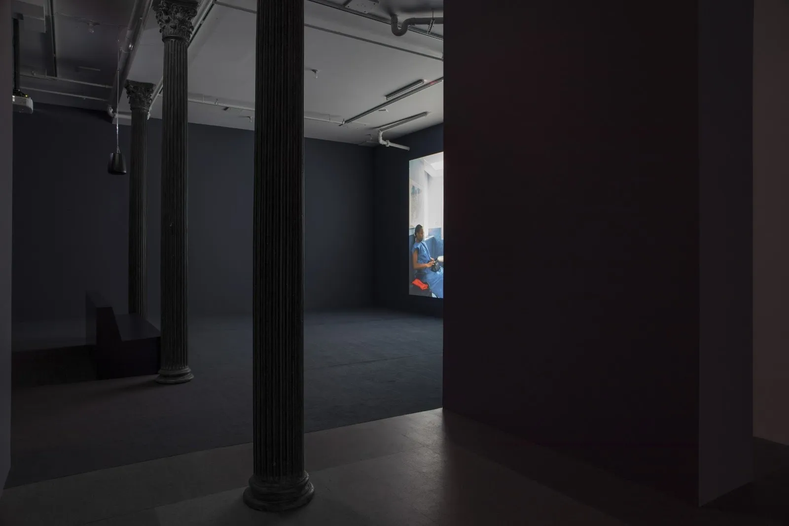 Shot of a dimly-lit gallery space with fluted columns. Partly obscured by another wall, a wall to the right of the shot projects a video.