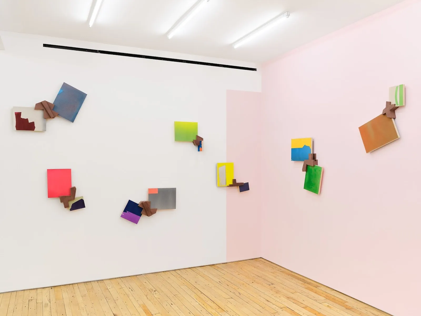 Installation view of wall-mounted abstract works in pastel-colored gallery space, featuring small geometric compositions combining painted panels and wooden elements.
