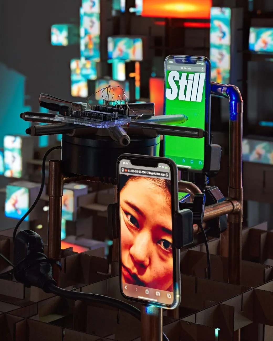 metal pipe structure supports various wires and two touchscreen phones. The phones display media, one displaying a face and the other displaying text that reads "still" over a green screen.