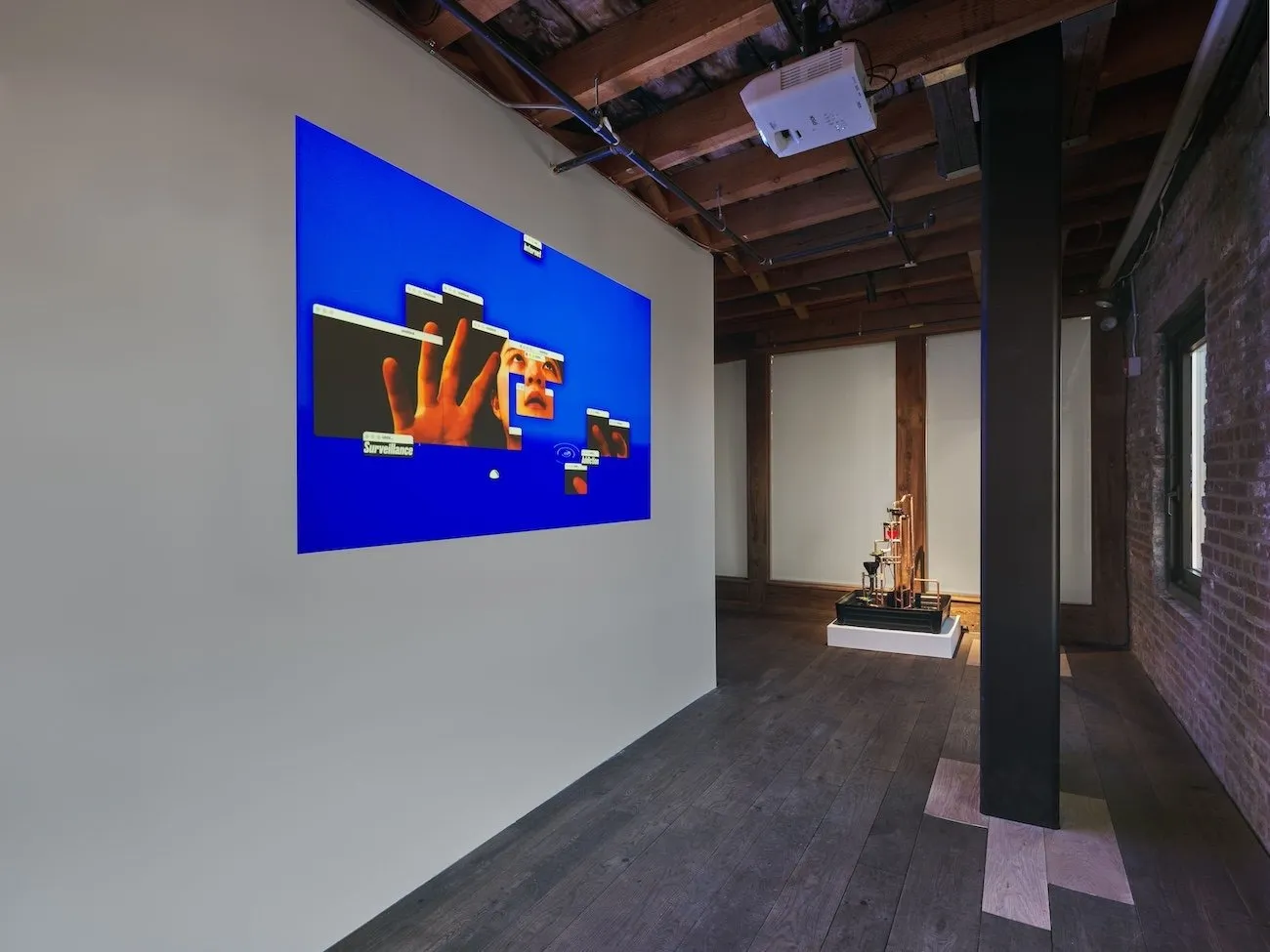Shot of gallery with hardwood floors, column, and beams. A bisecting white wall featuring a tv-sized projection displays a blue screen with various windows open, altogether displaying a face and hands.