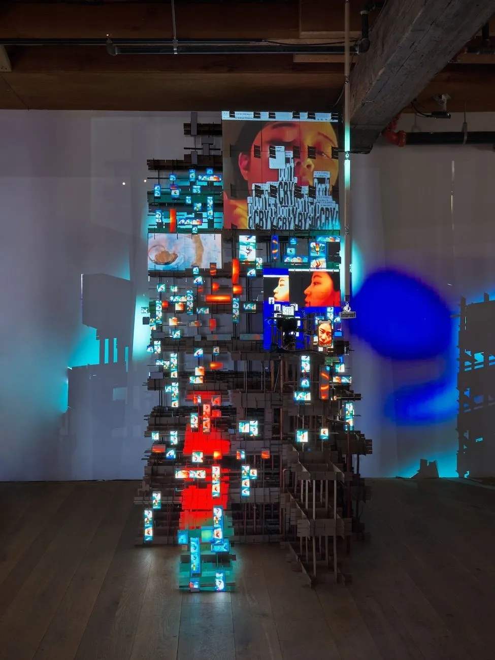 Gallery room with hardwood floors and beams and white walls. A scaffolding, stair-like sculpture supports multple screens of various sizes. Each screen displays media, including a face, text that reads "cry," and abstract imagery.
