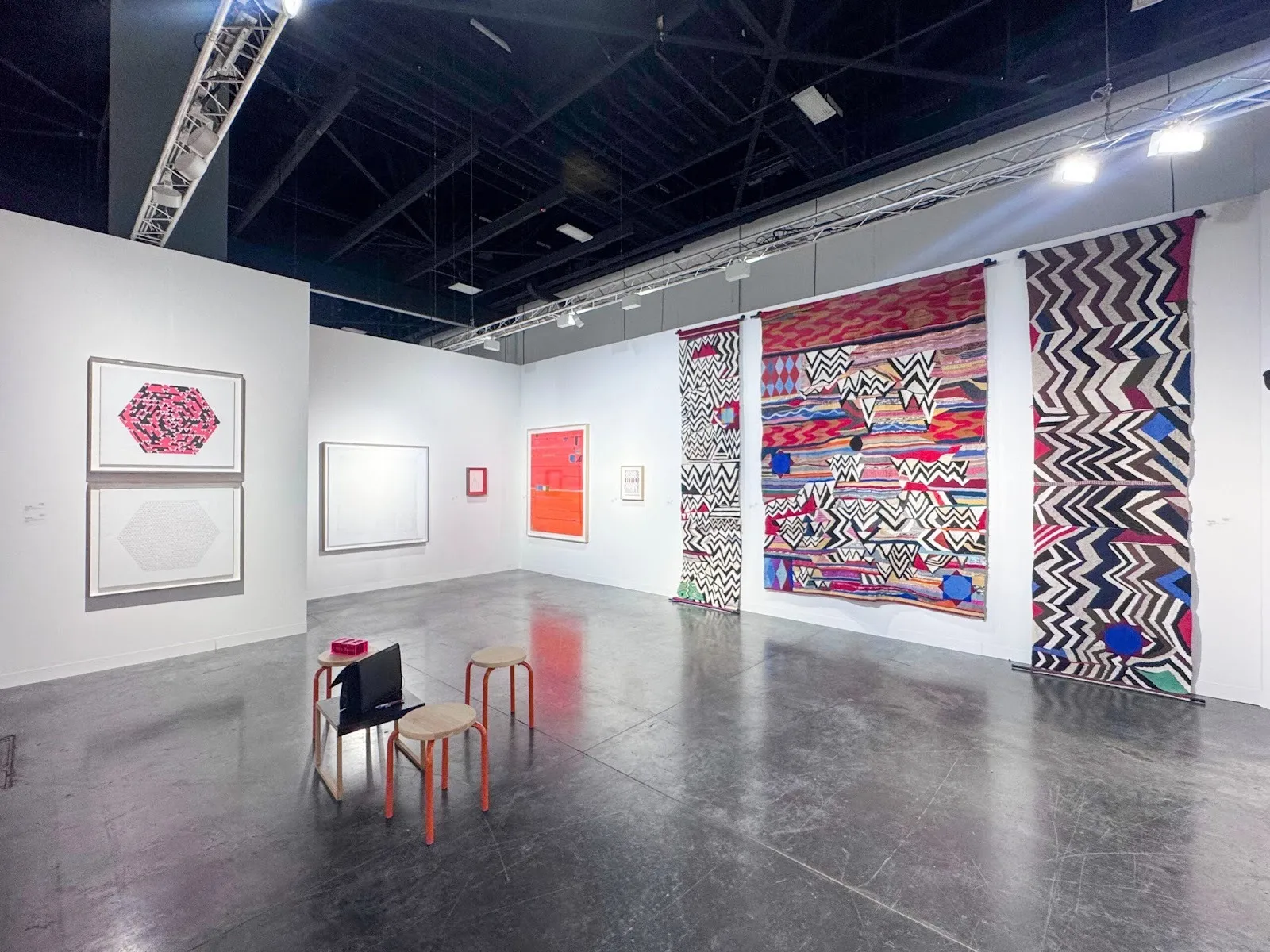 Art fair booth with framed geometric works and three large, colorful patterned textiles hanging on the right wall.