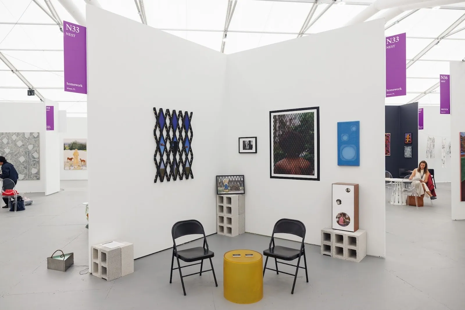 Corner booth with two black folding chairs, a yellow round stool, cinder blocks, and mixed media works on white walls under purple signage.