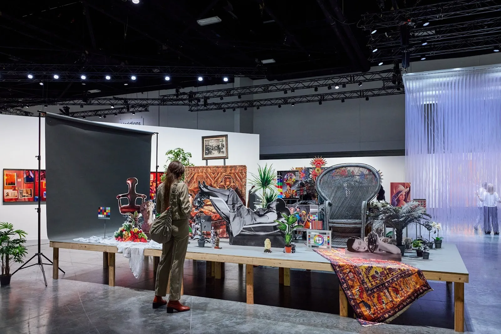 Large staged installation on a platform with a backdrop, cutout figures and furniture, plants, and patterned rugs, viewed by a person standing nearby.