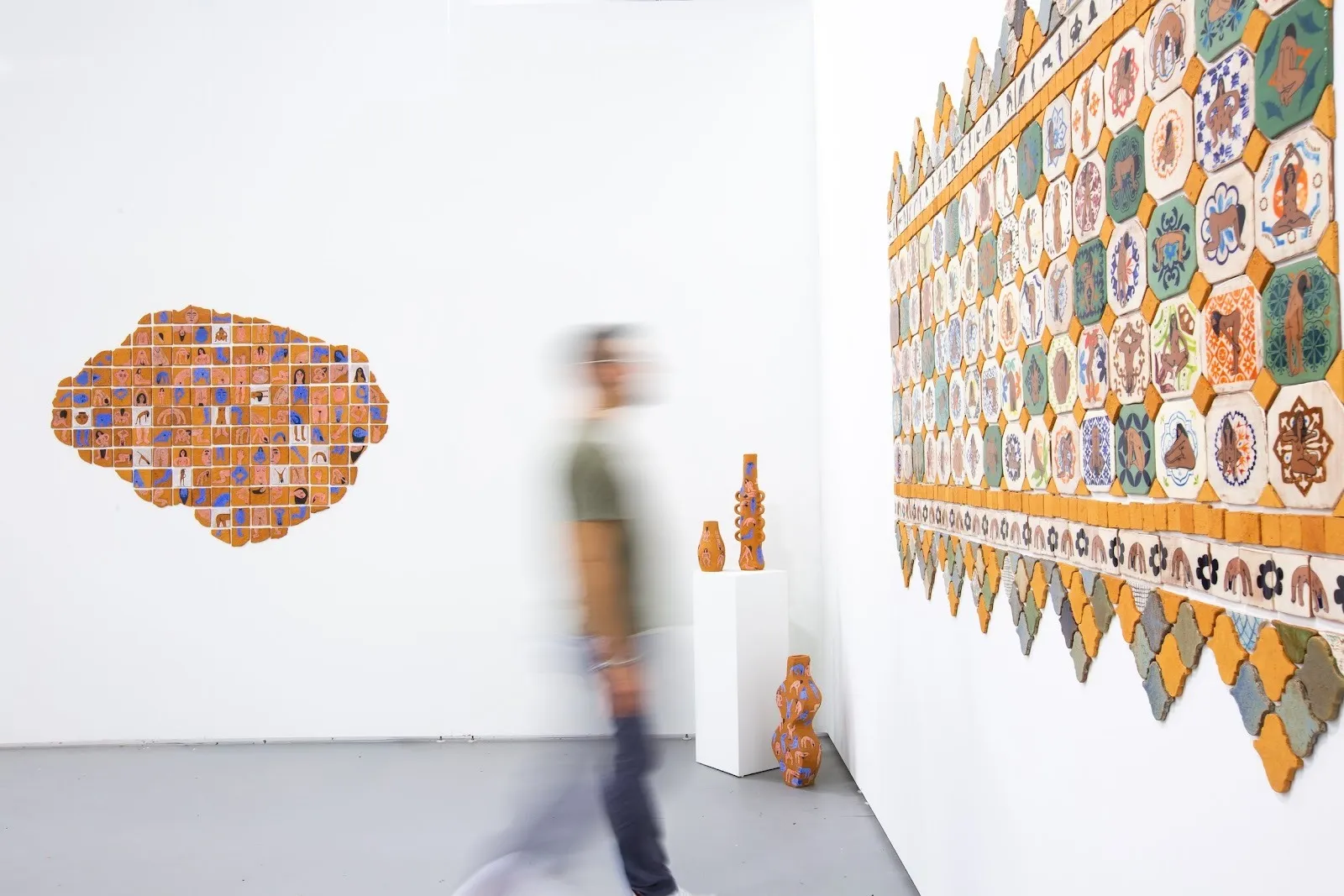 White gallery wall with orange-toned ceramic tile artworks arranged in geometric patterns; a blurred person walks past and small ceramic sculptures sit on a pedestal nearby.