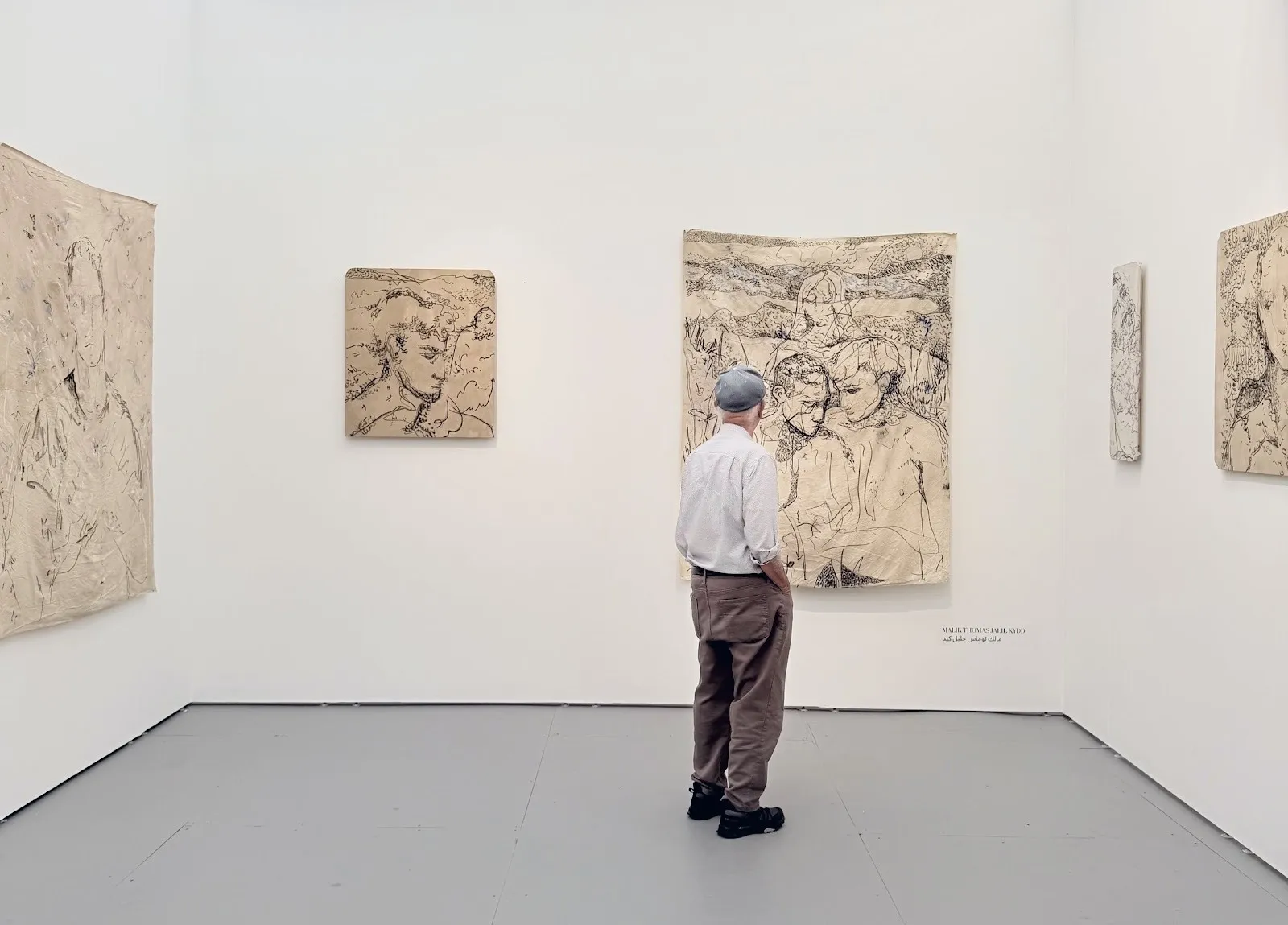 Man in a cap stands viewing monochrome line drawings on fabric-like hangings inside a white booth.