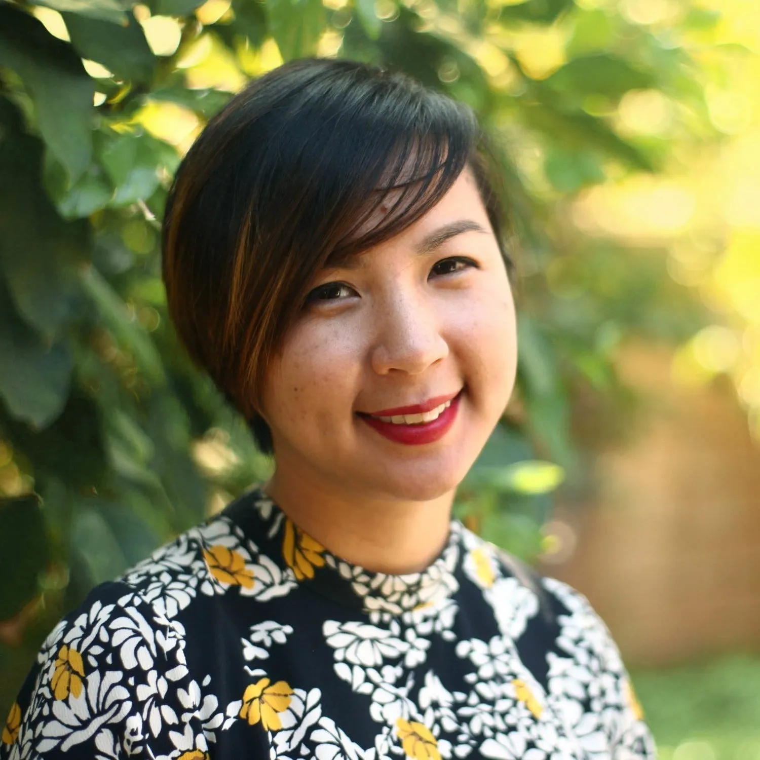 Headshot of a woman with a short hairdo smiling at the camera in front of a sunlit, leafy green background.