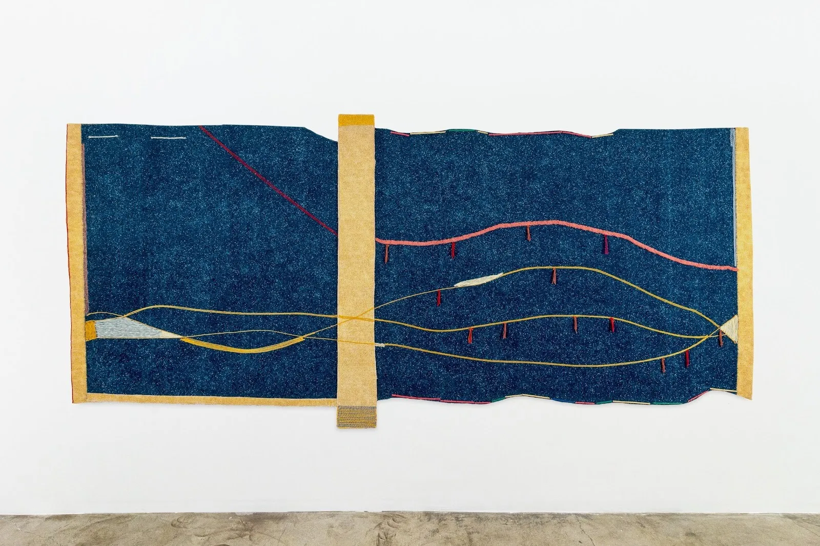 Shot of a large, horizontal rectangular work hung to a white gallery wall. The piece is irregularly shaped but features geometric, rectangular boxes of varying sizes. Dark blue astroturf is crossed over by colorful, meandering lines.