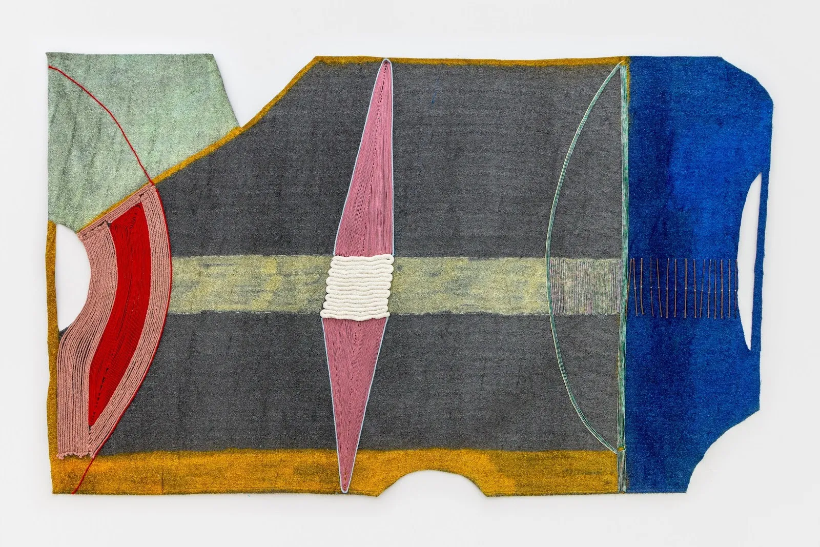 Shot of a horizontal work hung to a white gallery wall. The piece is 2D and vaguely rectangular-shaped, featuring geometric shapes like thin diamonds and half-circles of blue, grey, and red astroturf and thread. The sides have geometric, cut holes.