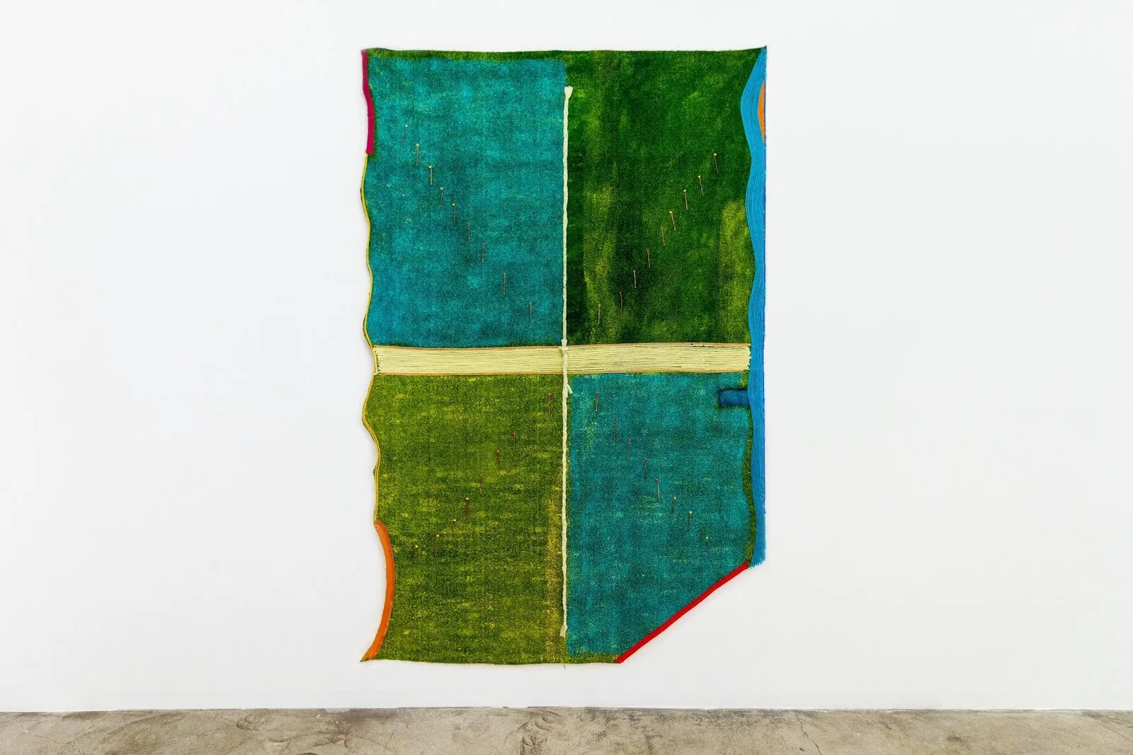 Shot of a large, rectangular hanging piece on a white gallery wall. The piece has both straight and rough edges and is designed into four spaces separated by a central cross. Each quadrant is either turquoise or green and made from artificial turf.