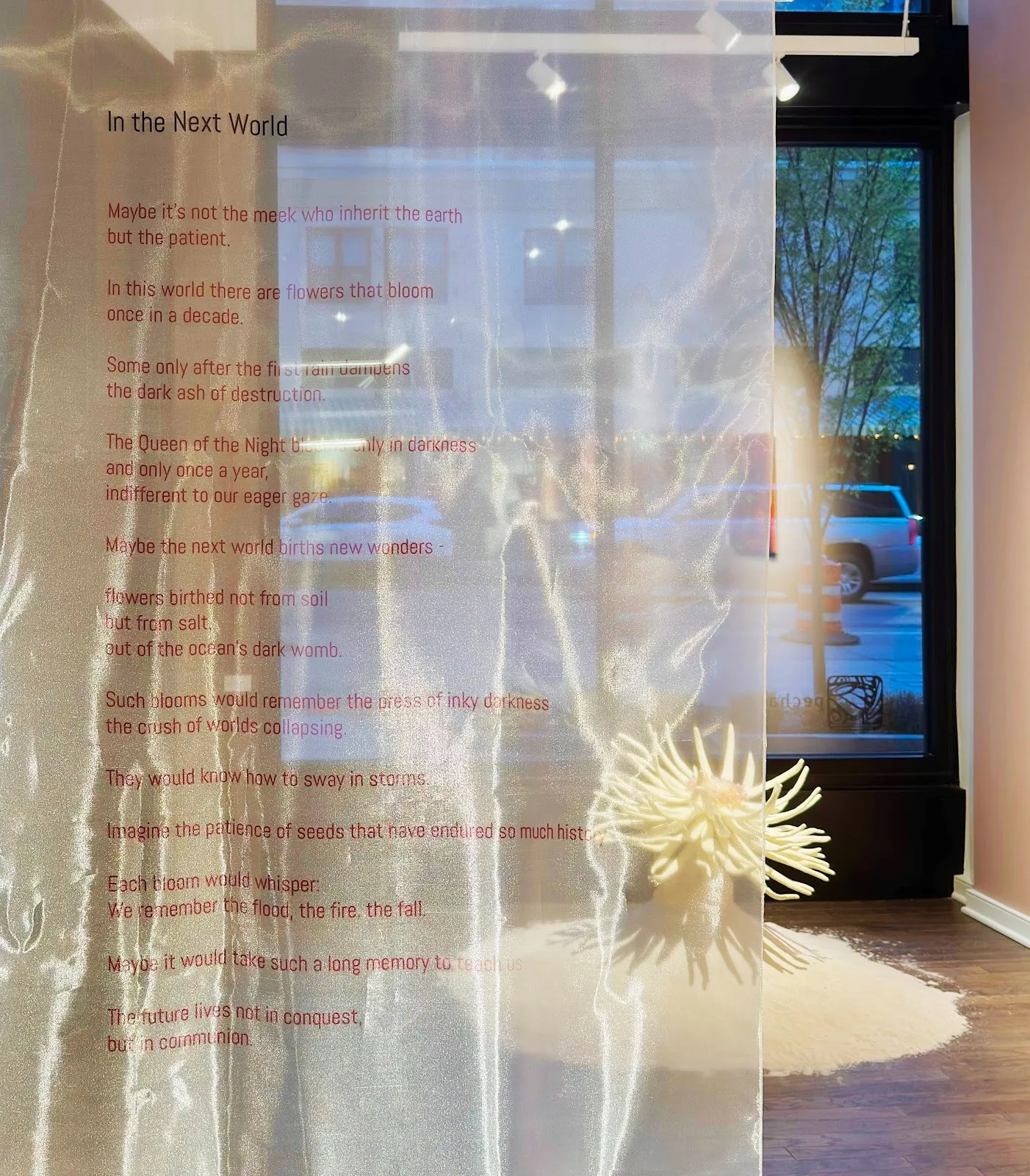 In a gallery space, a shiny, translucent swath of fabric hangs in the forefront. Red text is printed on the fabric, featuring a poem. In the background, a white flower rests on the floor and windows reveal a streetfront.