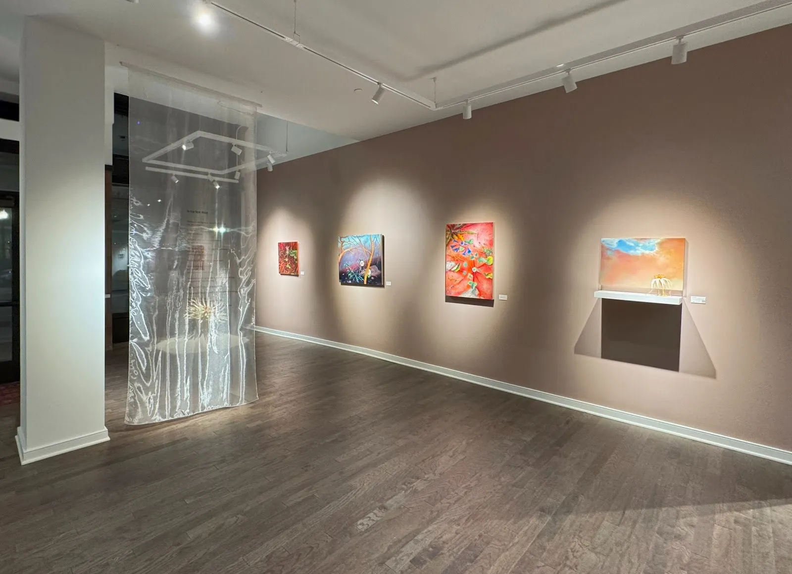 Shot of a gallery with beige walls and brownish hardwood flooring. Mutliple paintings of varying colors hang on the wall. To the right, a shiny, translucent sheet of fabric hangs from the ceiling to the floor and features red text.