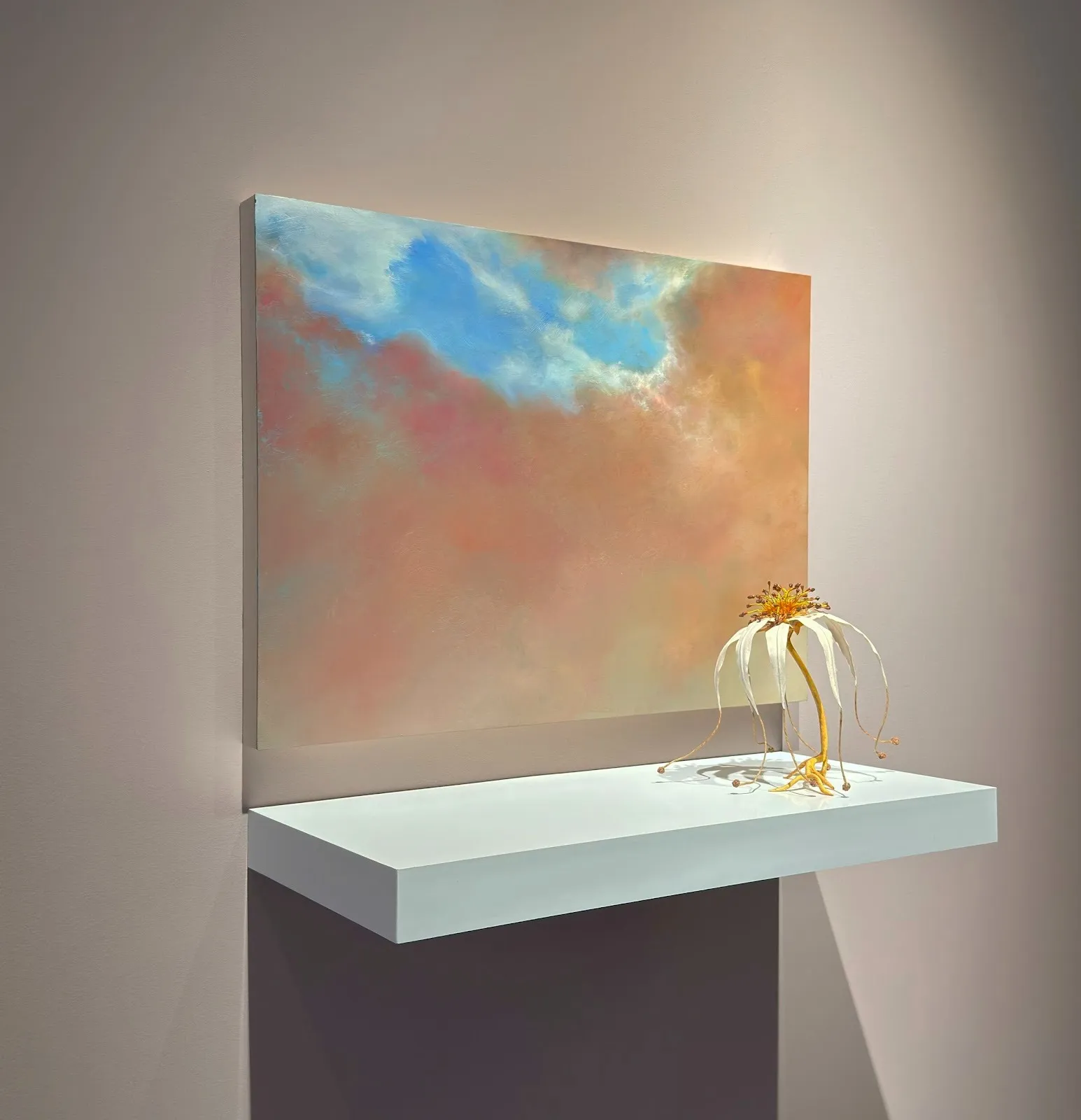 A medium-sized square painting of sky and clouds is mounted on a beige gallery wall and hangs over a white shelf of its width. On the shelf, a drooping, yellow and white flower rests to the right.