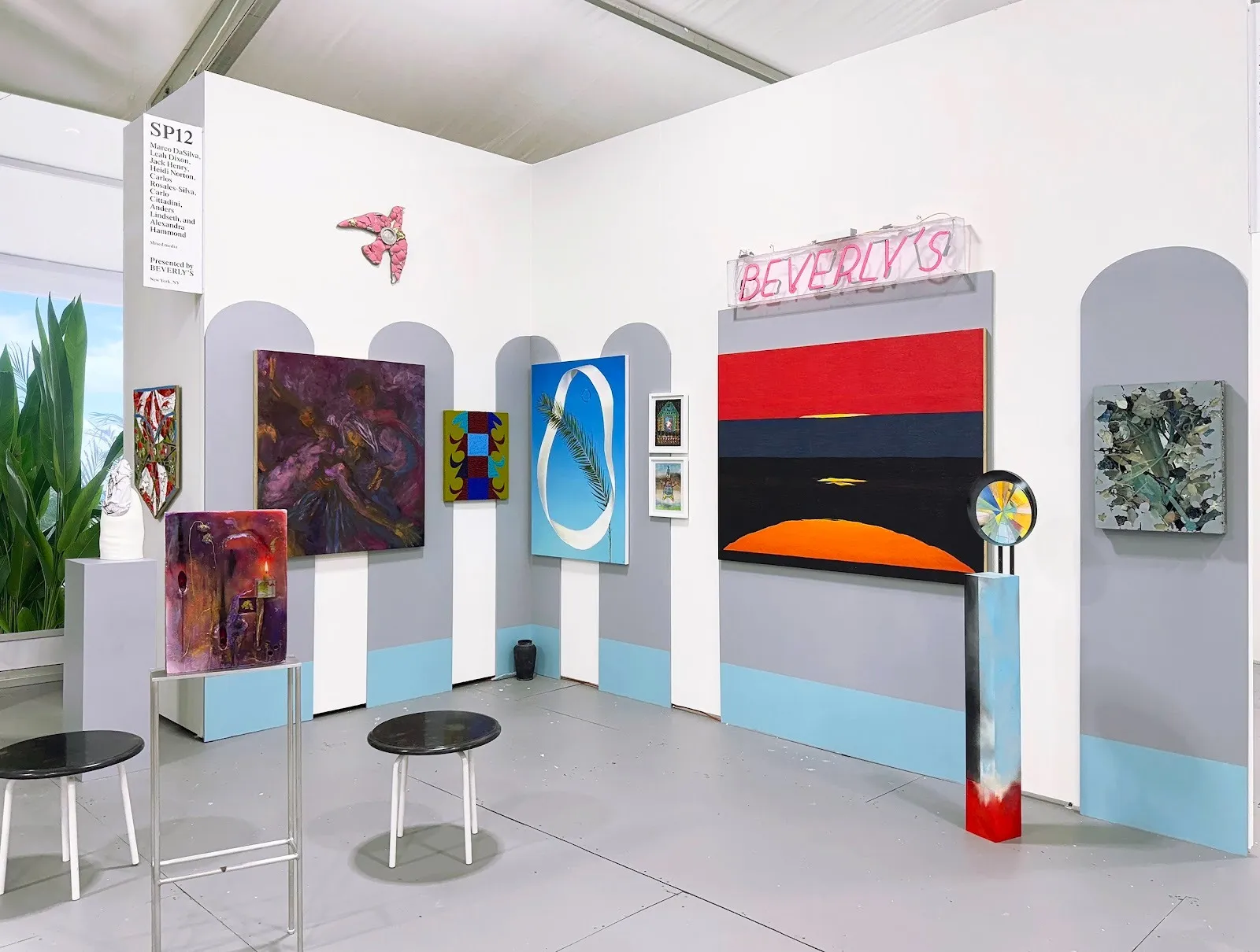 Booth with arched wall cutouts, a neon “BEVERLY’S” sign, and several paintings including a large striped sunset-like canvas.