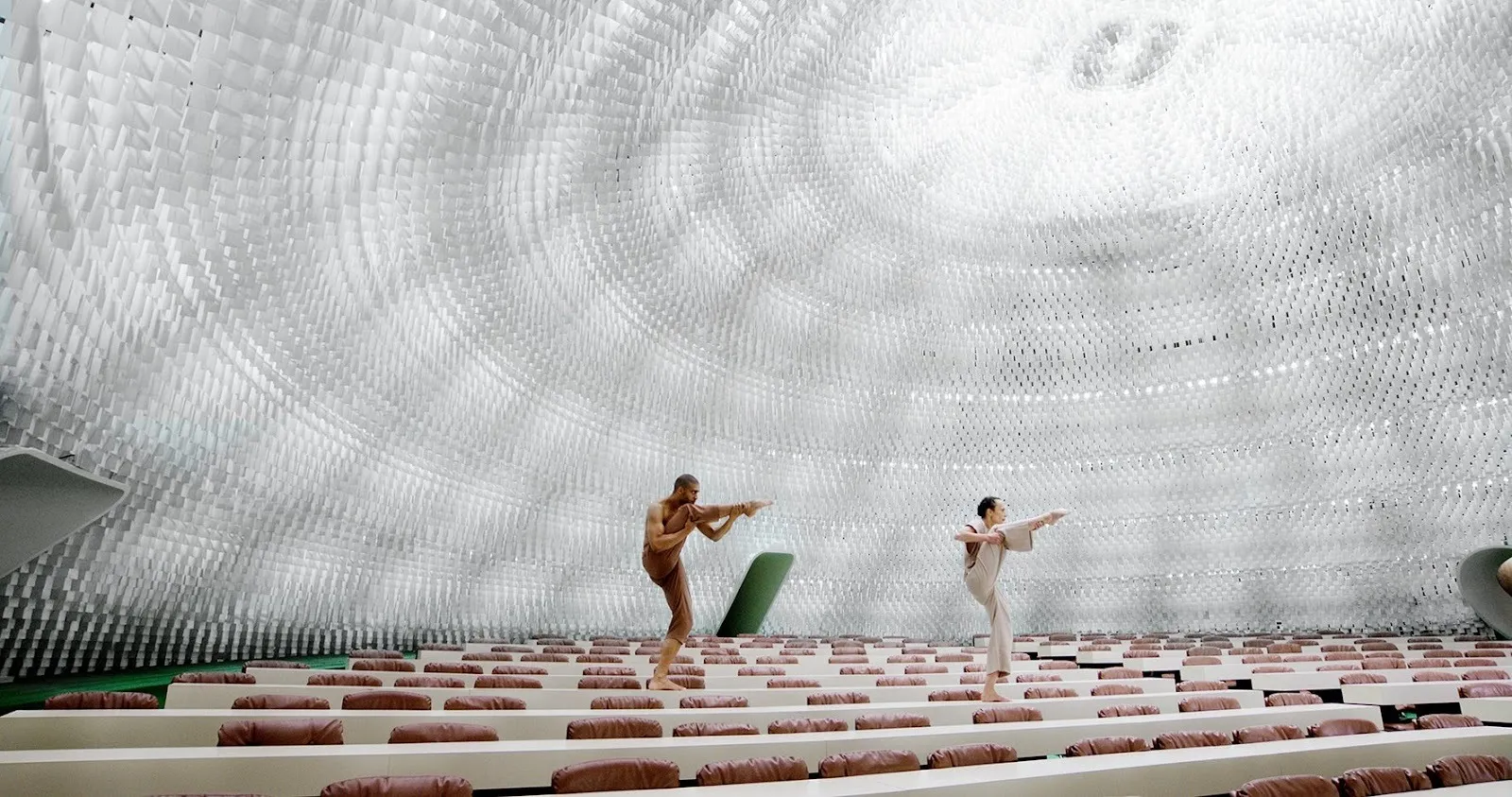Two dancers holding their legs up under a giant gray Dome, the gift by jasmina cibic.