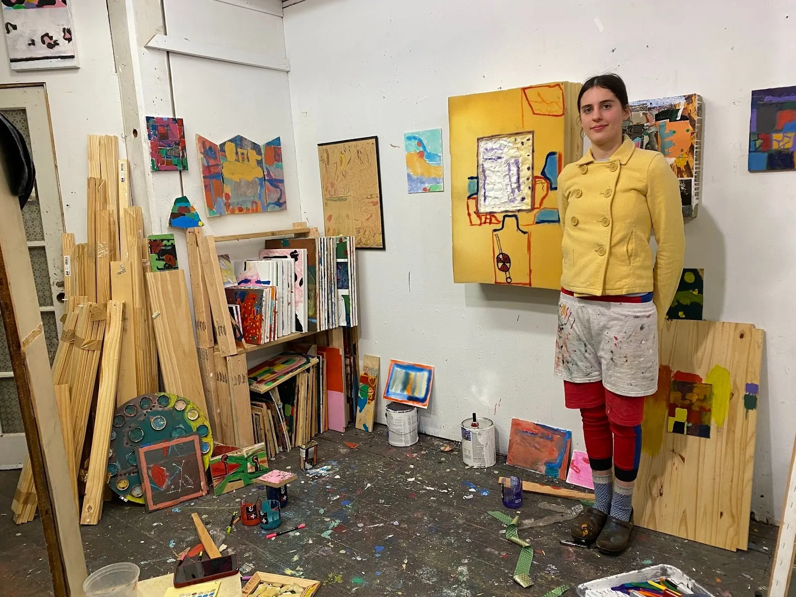 A white wall studio is filled with a paint-marked floor, wooden planks, paint cans, and various works mounted to the walls. To the right, a person wearing a yellow coat and layers of paints with paint stands, their arms behind their back.
