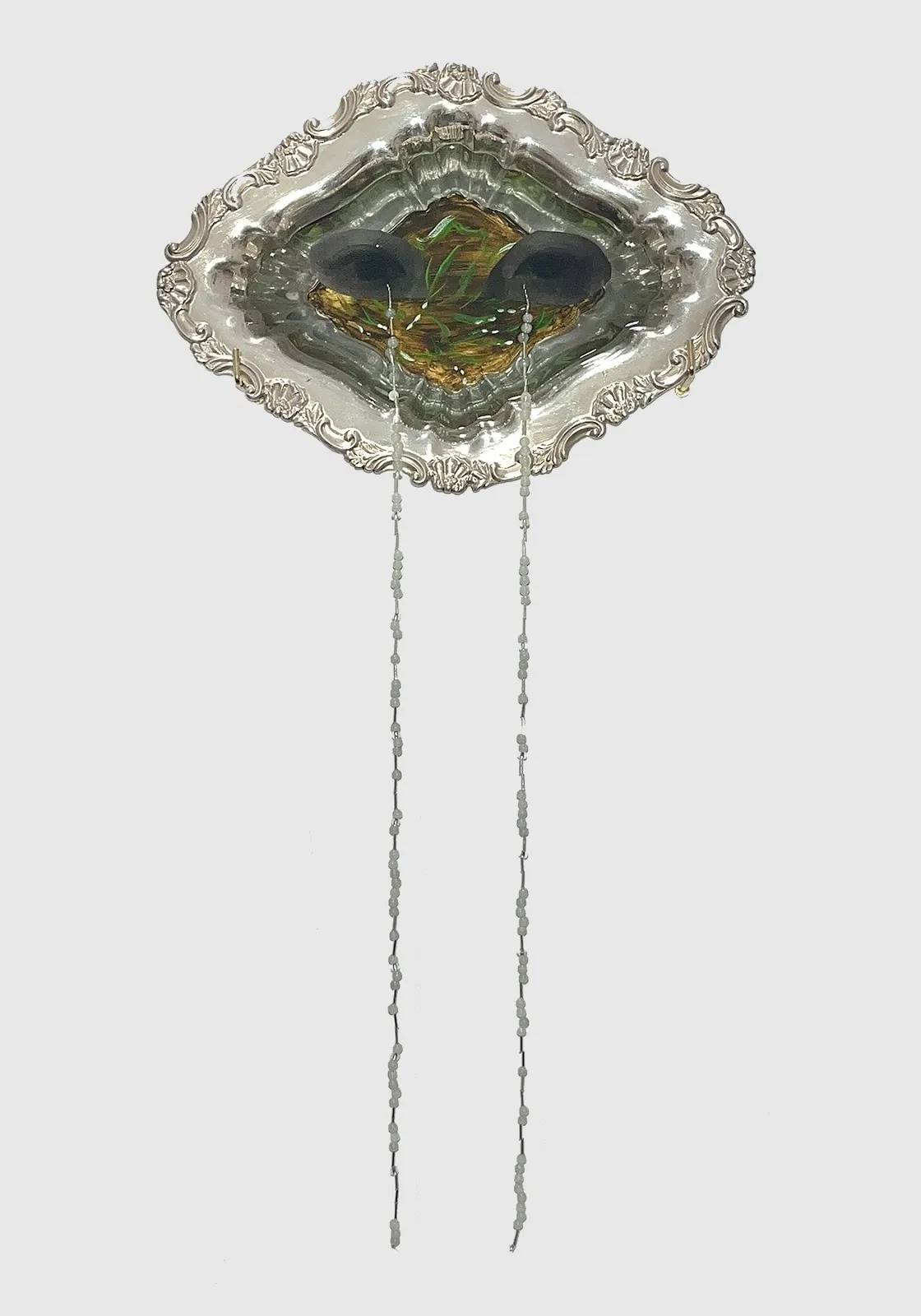 A decorated silver rhombus displays a painted surface within: eyes overlaid on flowering plants in the dirt. From the eyes, two long, thin strings of white beads hang down.