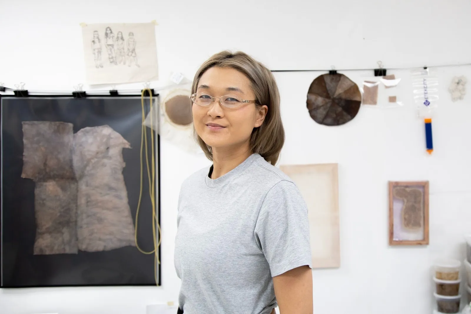 A person wearing a grey shirt and small gold glasses turns to the camera and smiles. Behind them, various works—biomaterials, sketches, objects—are hung to the white wall.