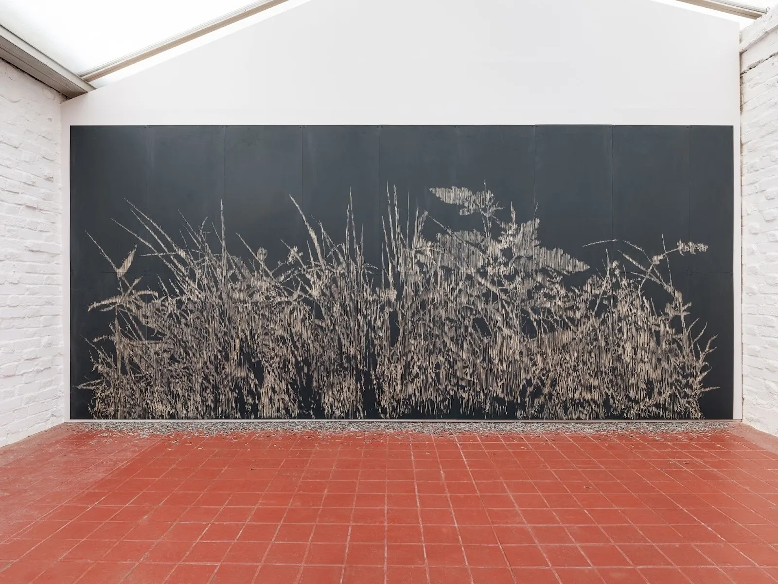 A black wall within a white structure bears marks that resemble a line of long grass or wheat. The terracotta tiled floor bears residue that resembles the grass.