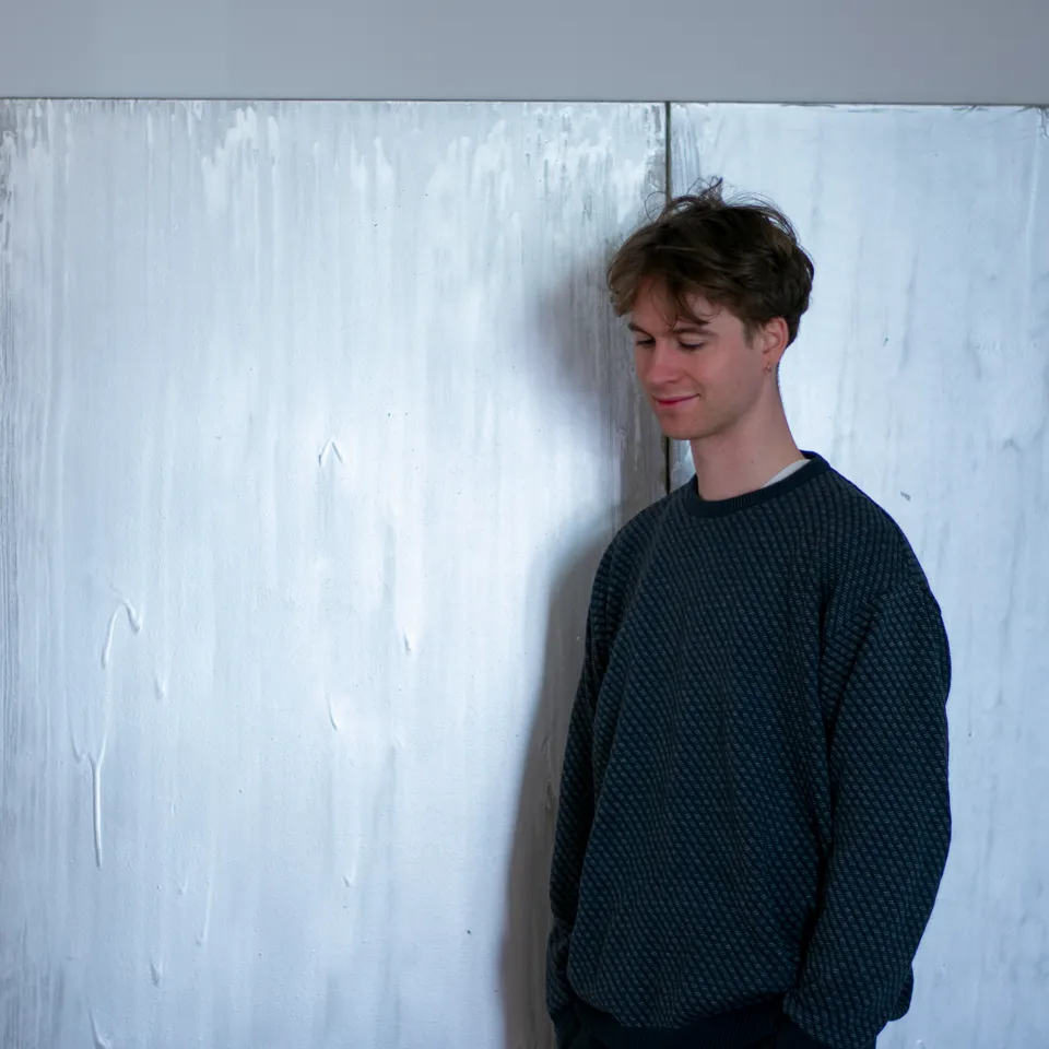 A person with short hair wearing a dark blue sweater stands in front of a large white painting. They look to the floor and smile.