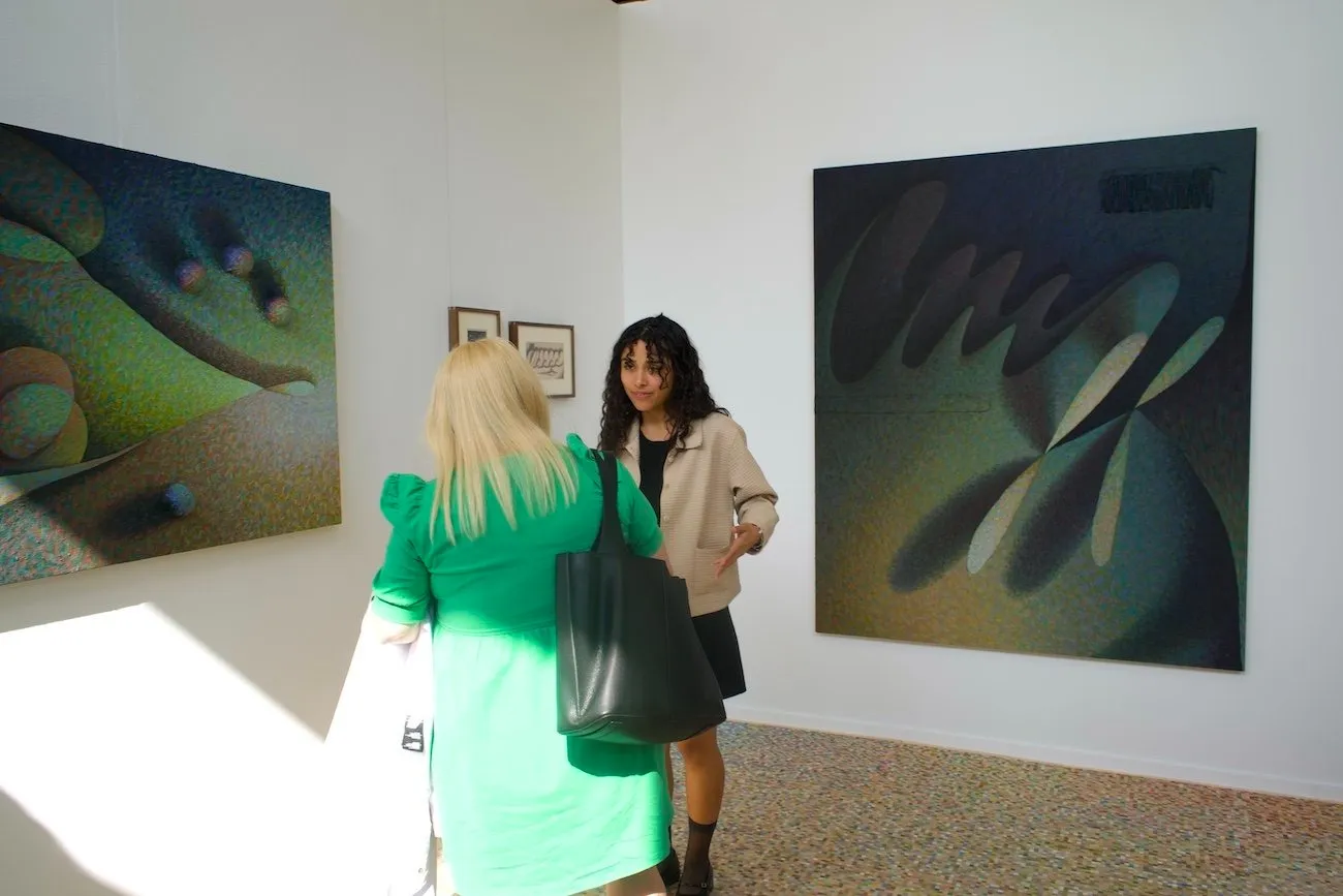 White gallery walls meet at a corner. Four works—two large unframed, two small framed, hang on the walls. The two large piece are done in a pointilist style, featuring abstract shapes and deep green/blue hues. Two figures stand and talk in front.