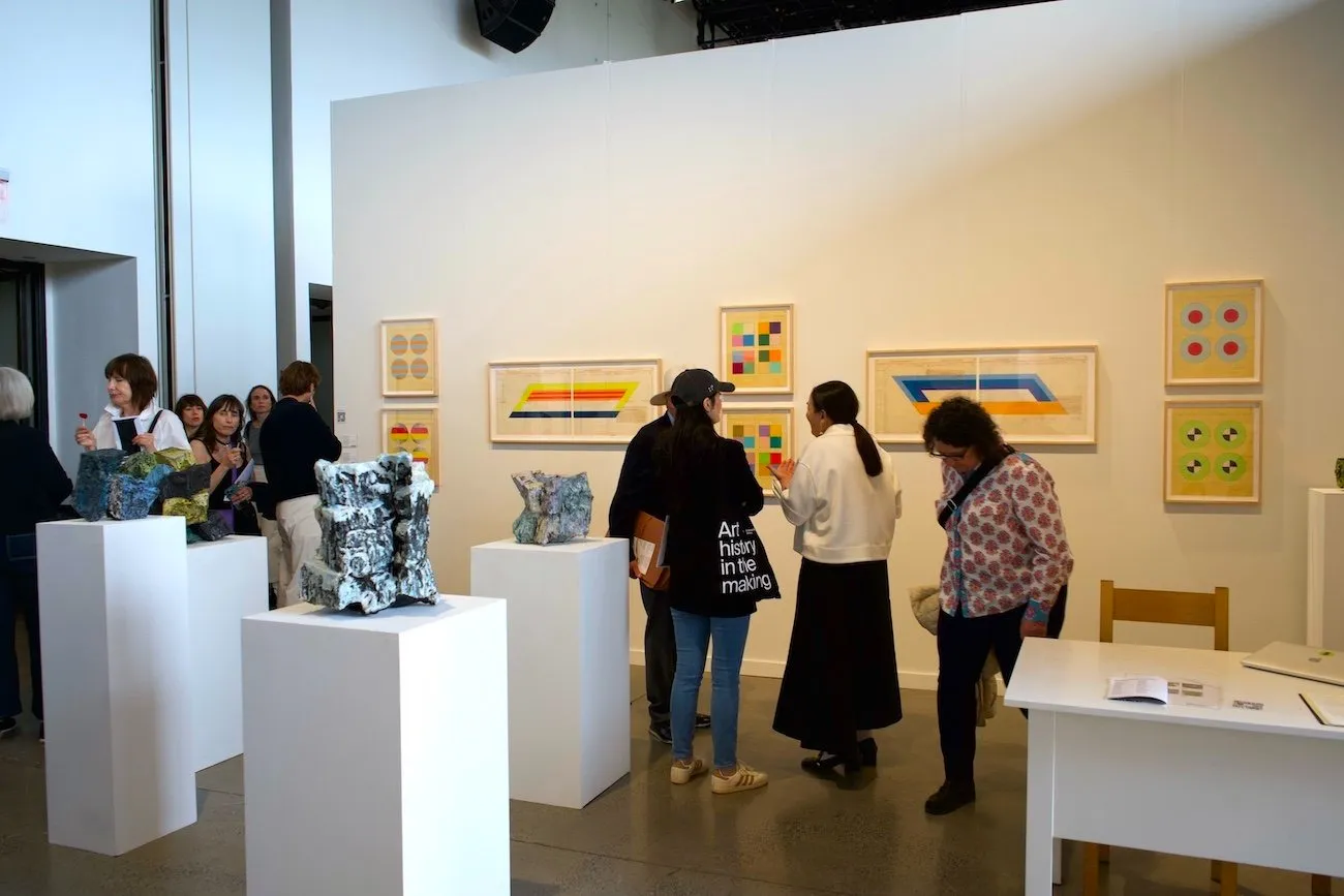 Multiple plinths and 2D pieces mounted to the wall scatter a white gallery space. The plinths feature abstract sculptures, while the wall works feature various colorful geometric patterns. Many people are scattered about the space.