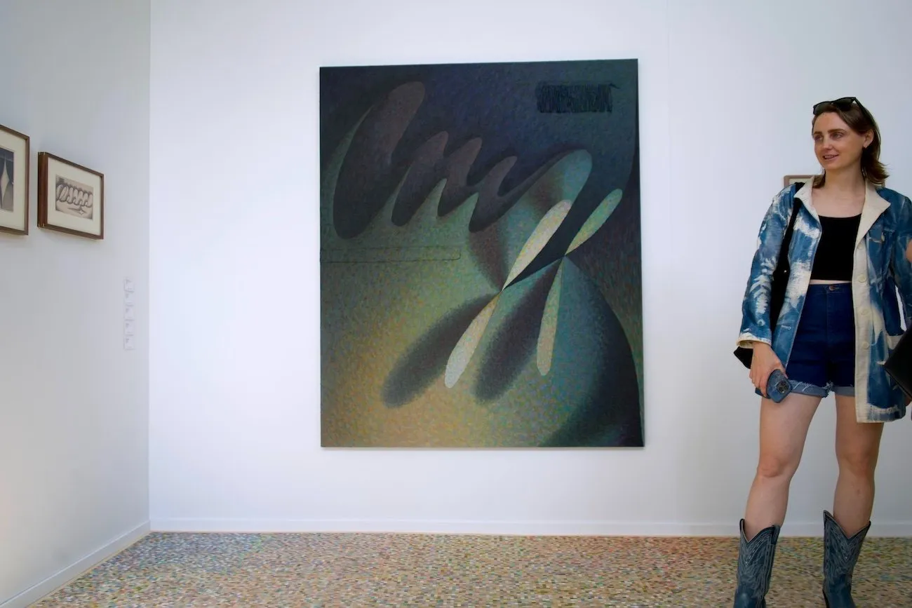 A person stands to the right of gallery walls displaying small and large works. The main large work is a pointilist abstraction made with deep green and blue shades.