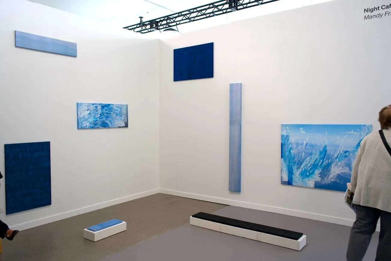 Two white gallery walls showcase a number of works of different widths and lengths. Two pieces lie on the floor. All pieces are different shades of blue and also differ in brushwork and texture.