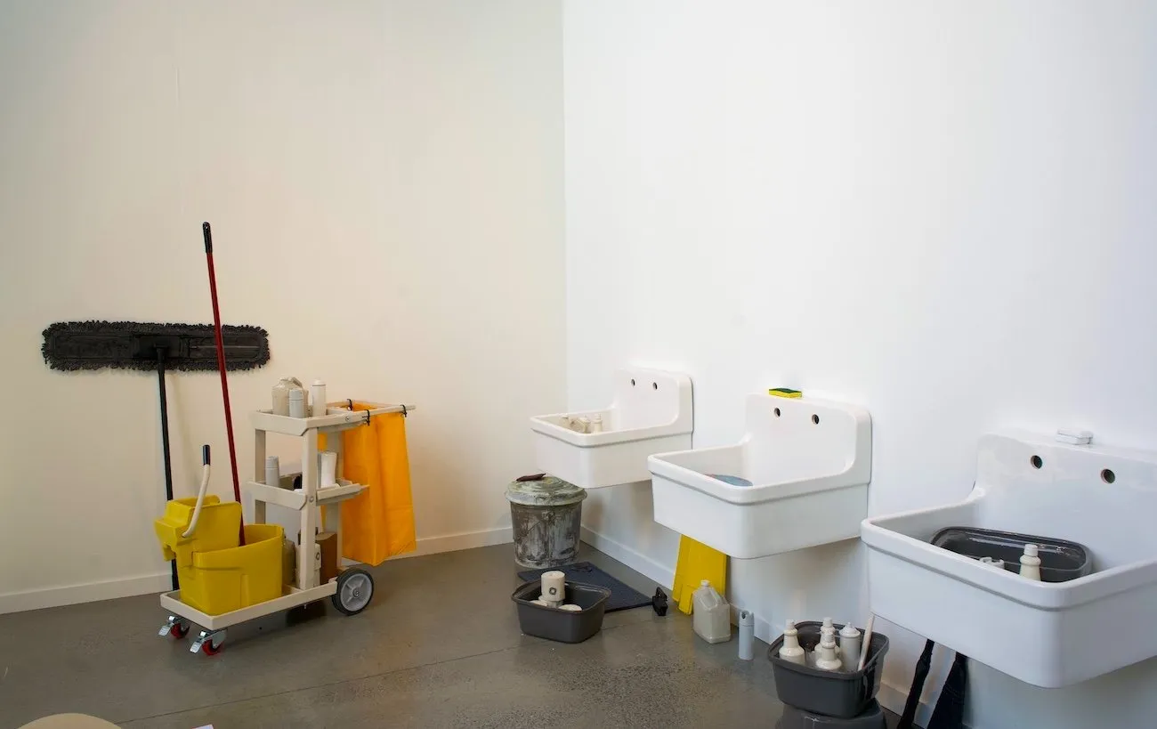 Three simple white sinks are mounted to a white gallery wall. Spread about the space are cleaning supplies and a custodial cart.