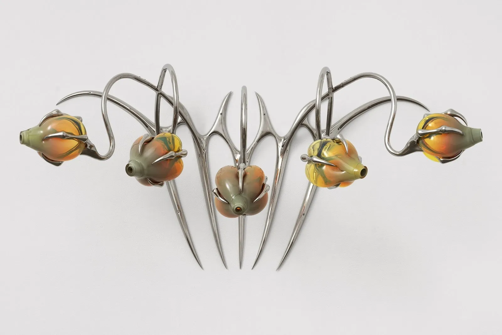 A symmetrical, sygil-esque piece made of silver metal which supports green and orange bulbous glass at its ends hangs.