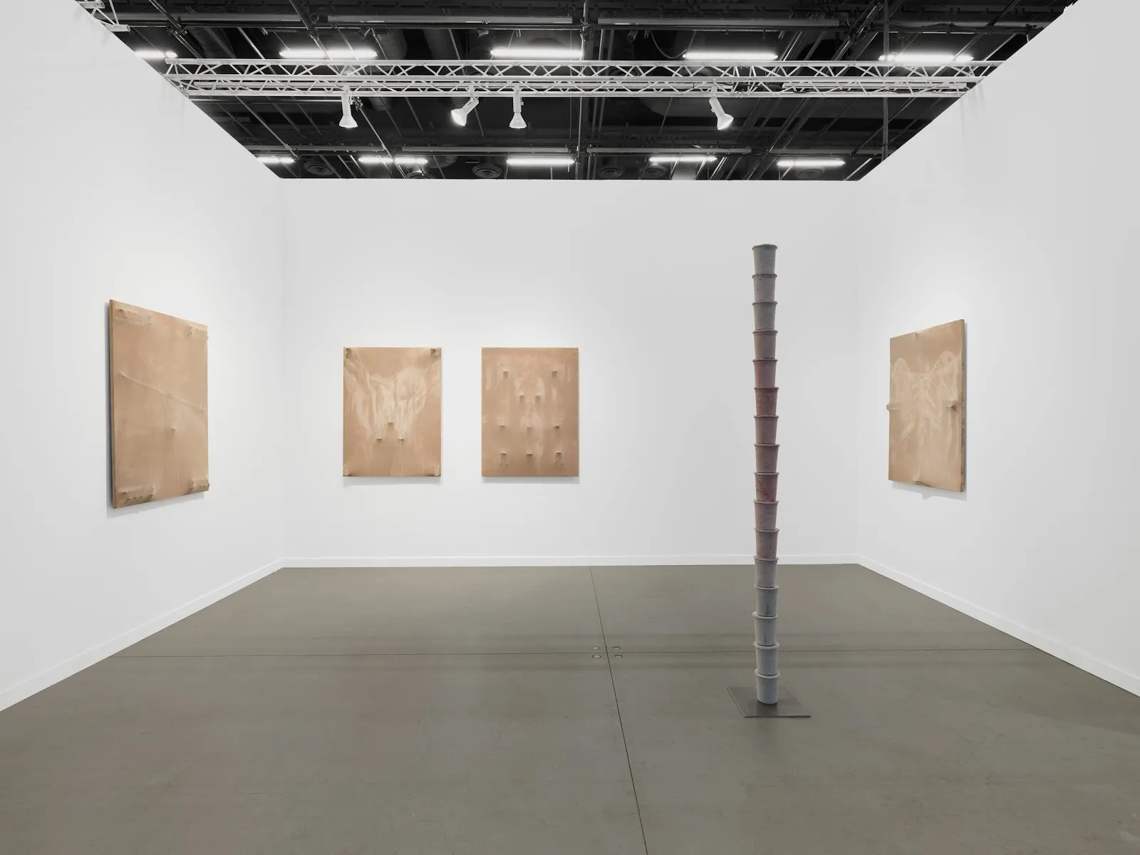 Three gallery walls support four 2D pieces and house one free-standing sculpture. The 2D pieces are all of similar sizes and the same tan shade. The sculpture is grey and tall, akin to stacked cups.