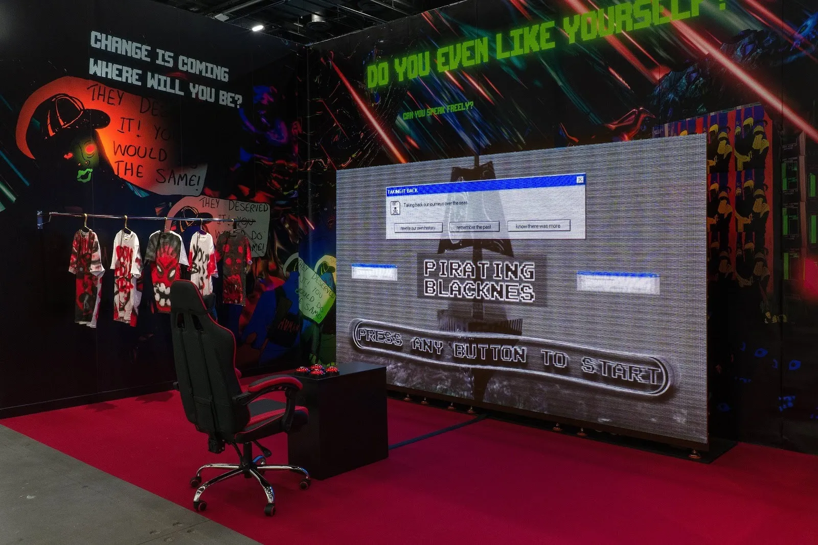 Gallery walls feature art and text from floor to ceiling. In the space, red carpeting, a rack of clothes, a gaming chair and console, and a large screen stand.