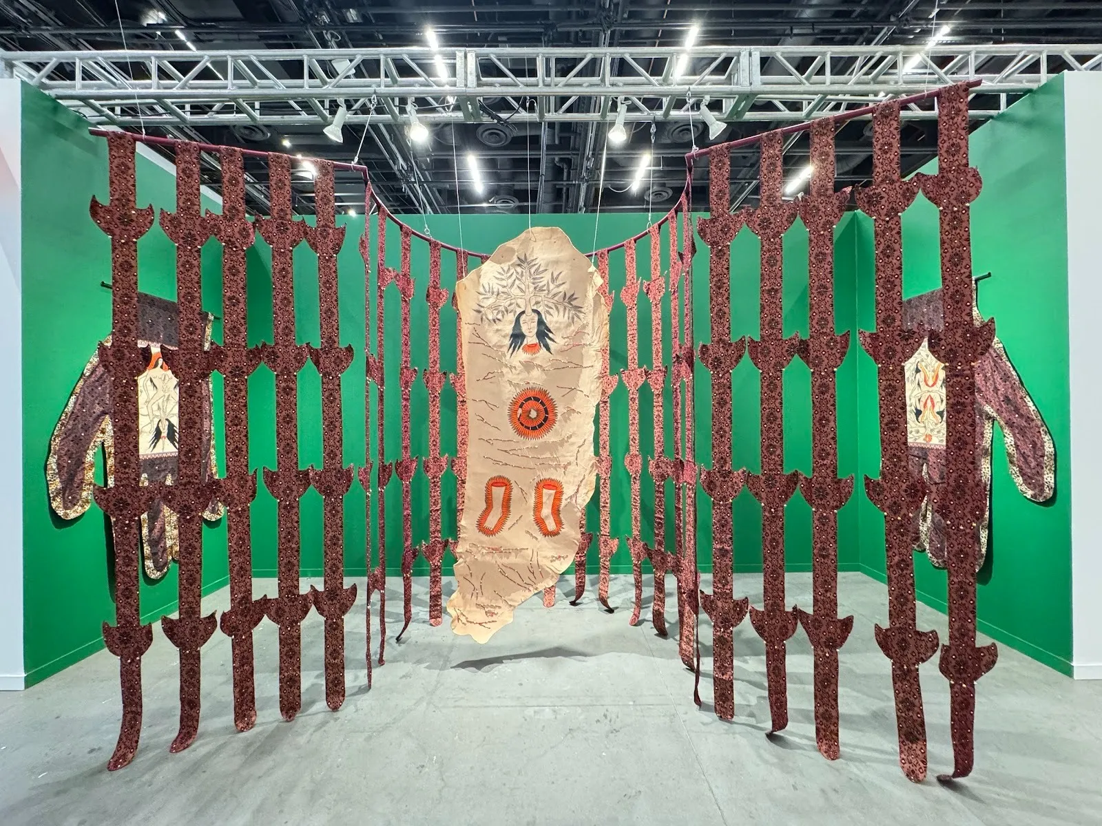 Three green walls stand below warehouse-like scaffolding, which supports a hanging piece made from a fabric-like material. The fabric is hung in grid-like strips and features a central piece of an abstracted figure. Other similar motifs hang on walls