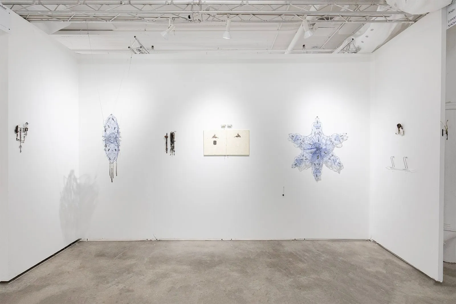 Three white walls support a variety of hanging and mounted works, all of different material, shape, and form. Some are translucent blue shapes, others are flat canvases, and others have amorphous, wiry forms.