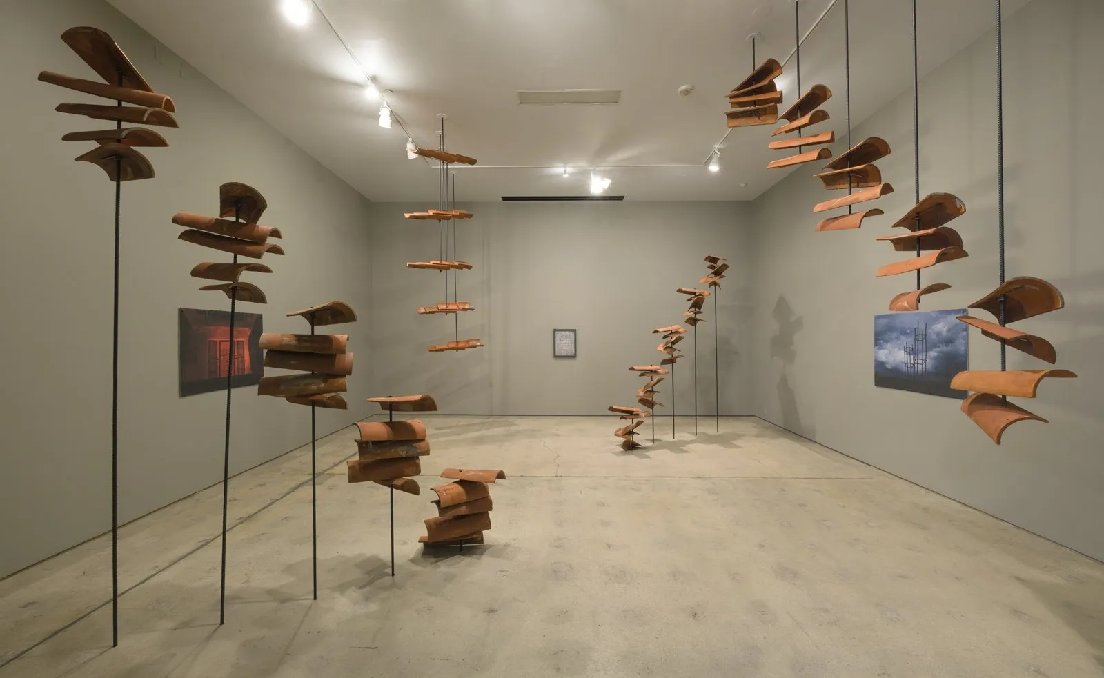 A grey room holds multiple hanging and supported sculptures made from roof tiles. Three medium-sized paintings are hung on the walls.