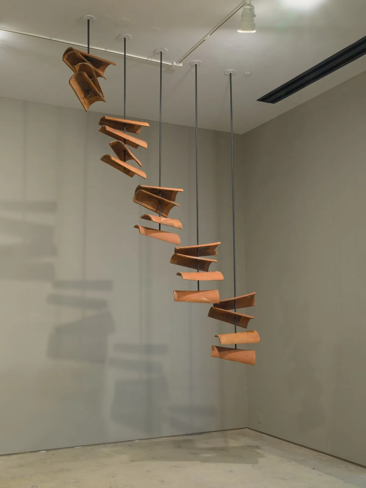 A hanging sculpture made from a row increasing lengths of rebars which pierce terracotta roof tiles.